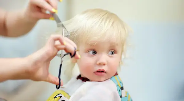 Un adorable petit garçon de 1 an, aux cheveux blonds, reçoit sa première coiffure. Ses grands yeux bleus regardent avec curiosité les ciseaux et le peigne.
