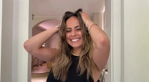 Une femme souriante avec des cheveux châtains ondulés, elle joue avec ses cheveux pour boucler cheveux. Elle porte des bracelets dorés.