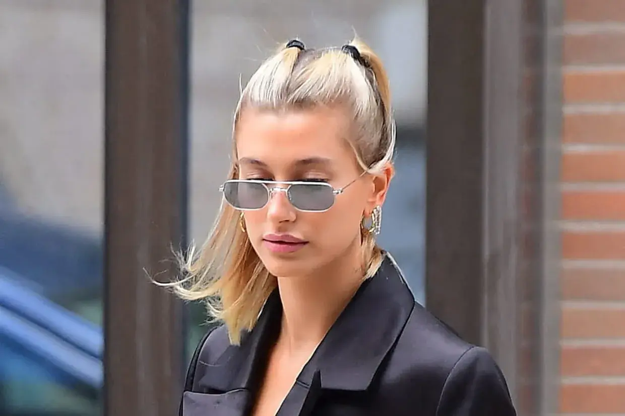 Une jeune femme aux cheveux blonds coiffés en deux couettes, portant des lunettes de soleil rectangulaires et une veste noire.