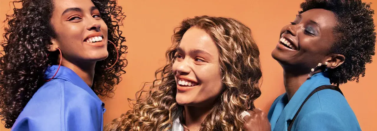 Trois femmes souriantes aux cheveux bouclés, chacune avec un style unique. Une inspiration pour savoir quelle frange pour quel visage.
