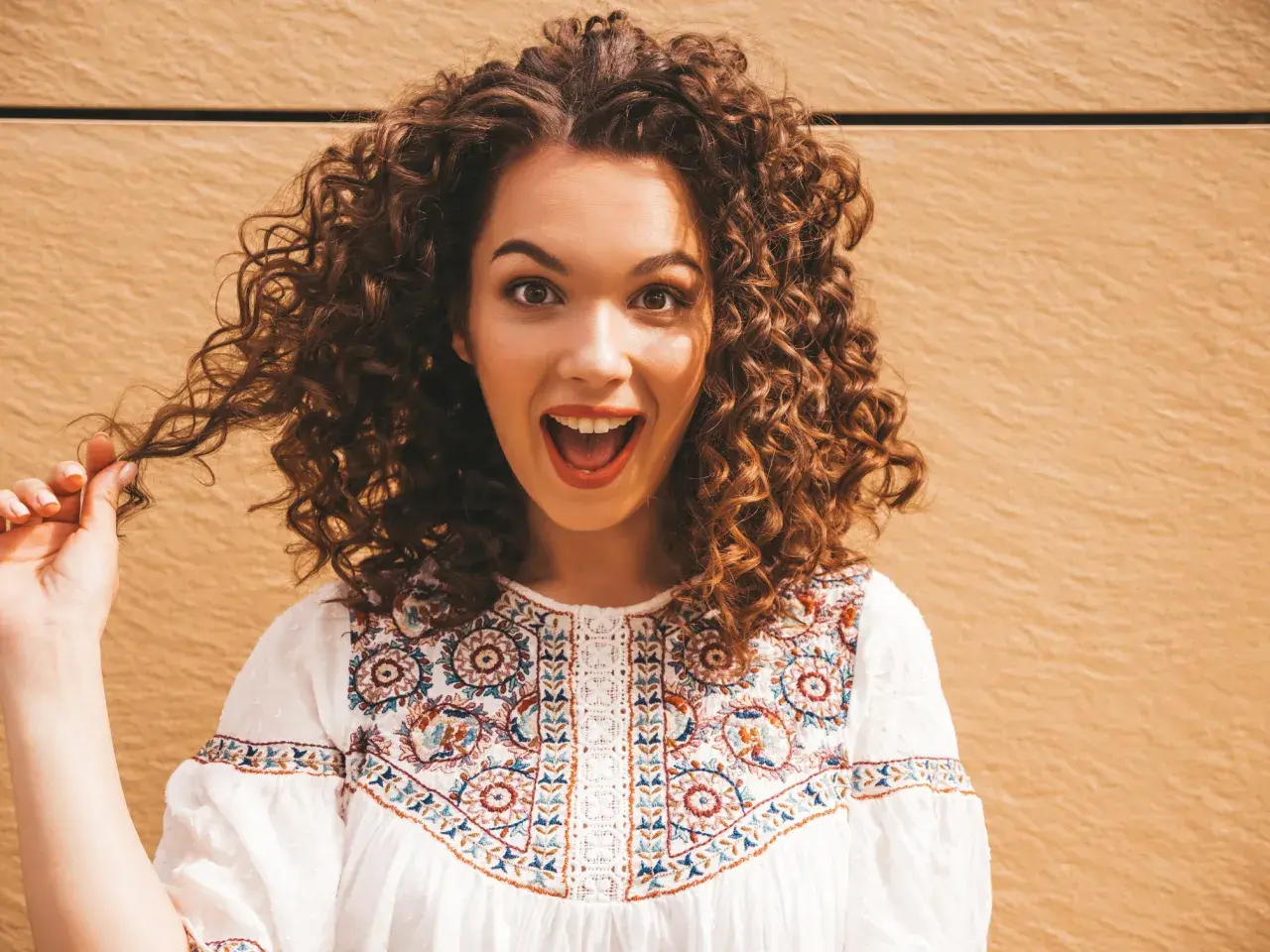 Une jeune femme surprise, sa coiffure cheveux bouclés tombant en cascade. Elle porte une blouse blanche brodée.