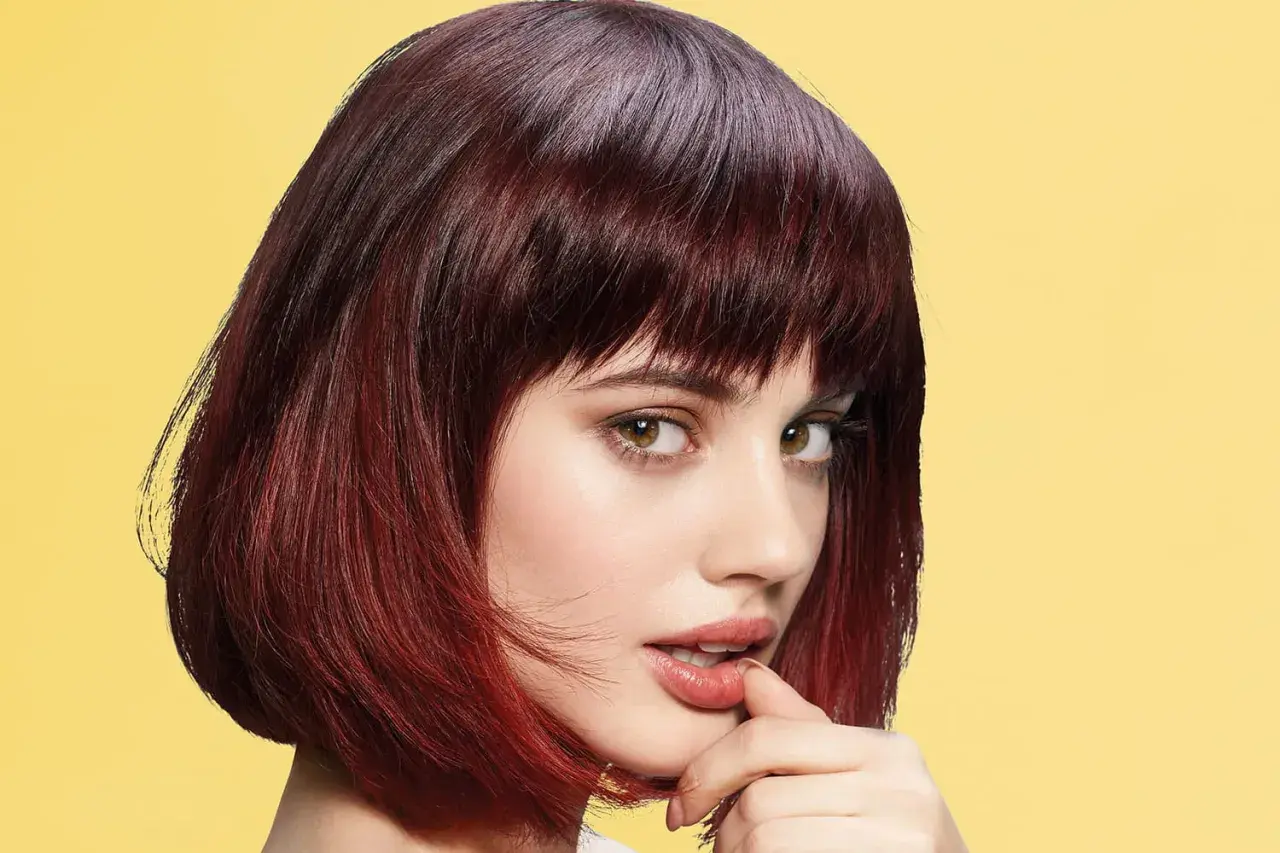 Une jeune femme au regard intense arbore une coupe cheveux epais, mi-longue, dégradée et colorée de reflets auburn sur fond jaune vif.