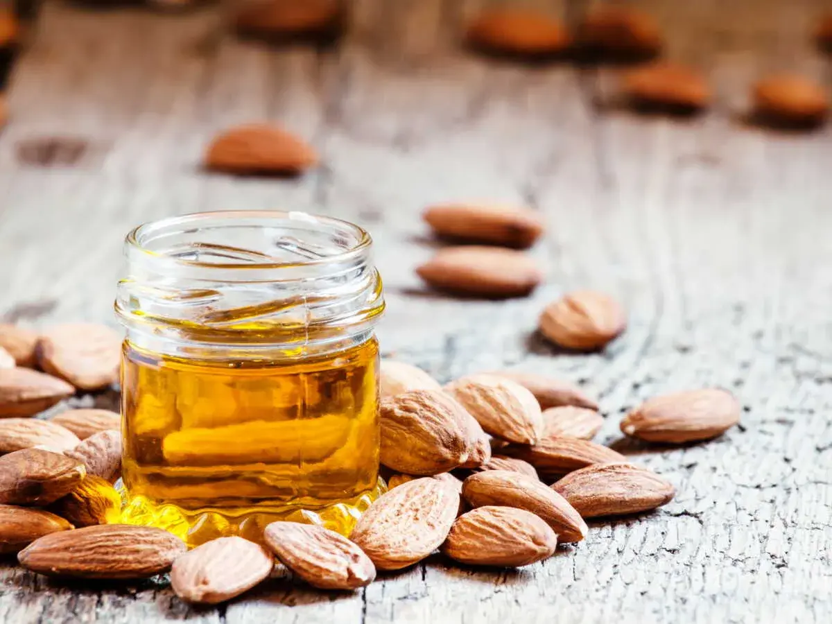 Huile d'amande dorée dans un petit pot, entourée d'amandes. Un secret naturel pour le bienfait des amandes sur les cheveux.