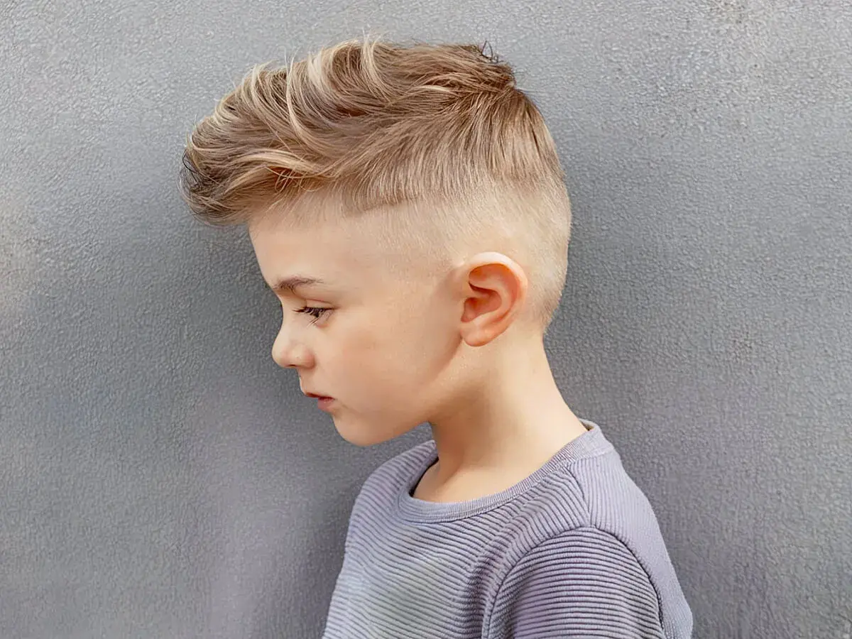 Un jeune garçon de 8 ans arbore une coupe de cheveux moderne avec un dégradé sur les côtés et des mèches blondes texturées sur le dessus.
