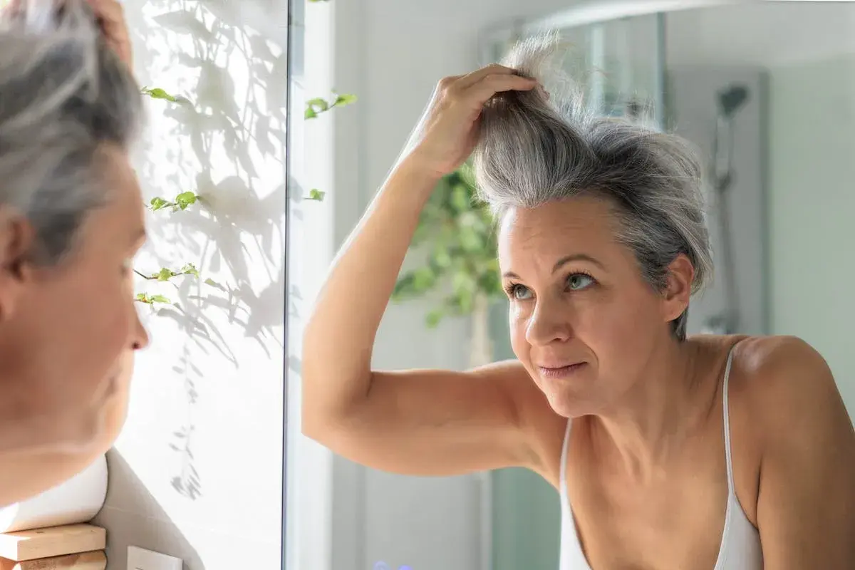 Une femme regarde ses cheveux poivre et sel dans le miroir, se demandant comment les blanchir pour un look plus lumineux.