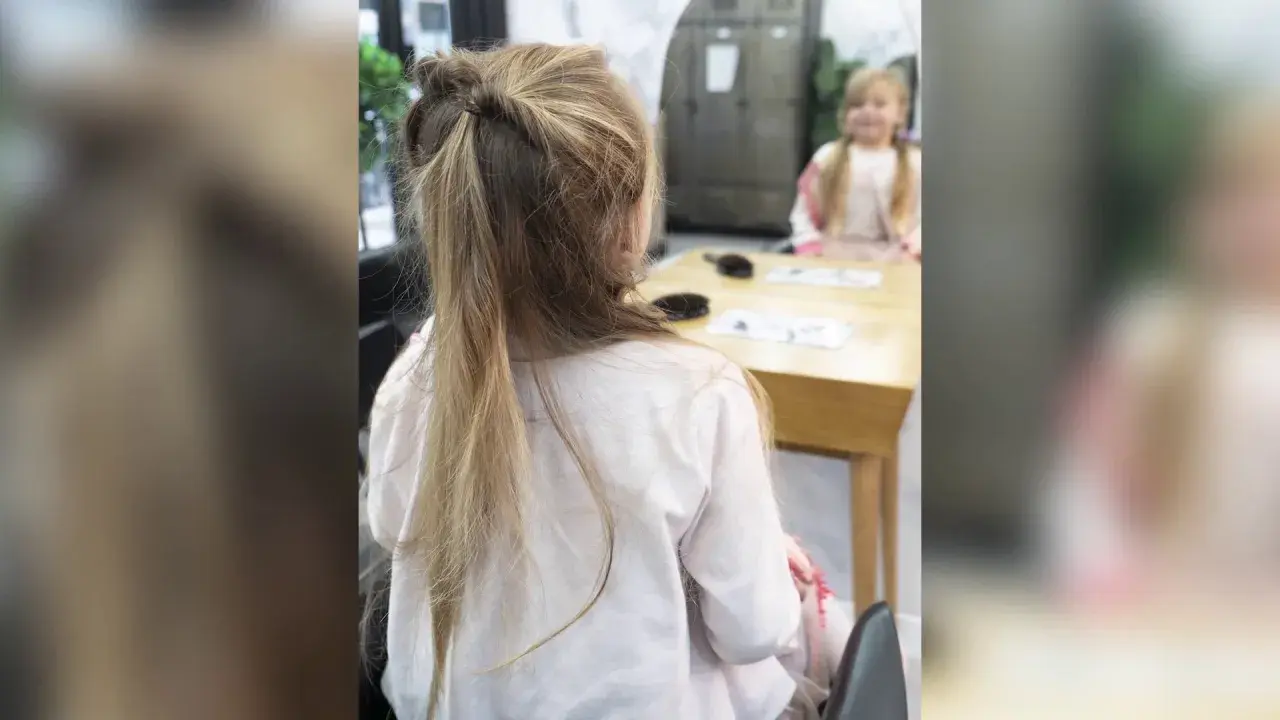 Une petite fille avec une jolie demi queue de cheval torsadée, assise à une table.