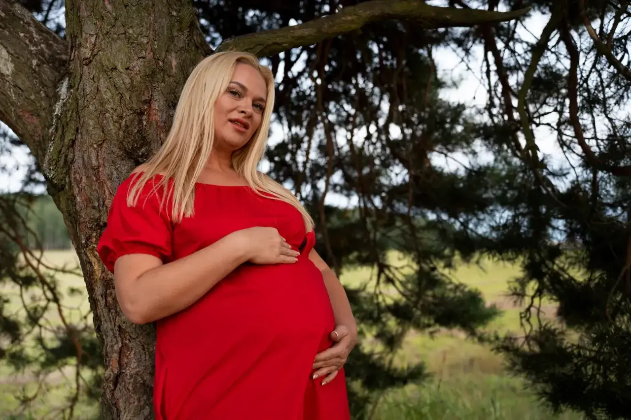 Femme enceinte en robe rouge, appuyée contre un arbre. Elle caresse son ventre, son regard est tourné vers l'objectif. On se demande si faire des mèches enceinte est dangereux.