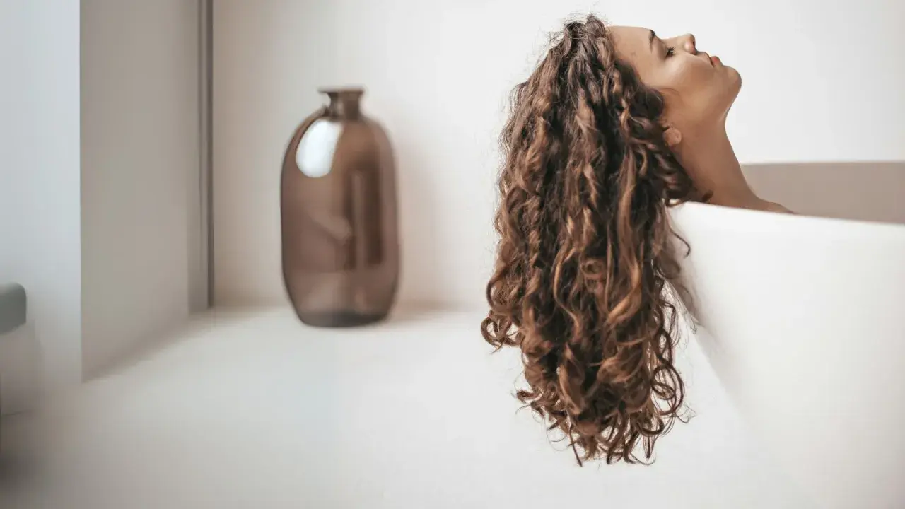 Une femme aux cheveux bouclés se détend dans une baignoire. Apprenez comment utiliser un après-shampoing pour des boucles douces et brillantes.