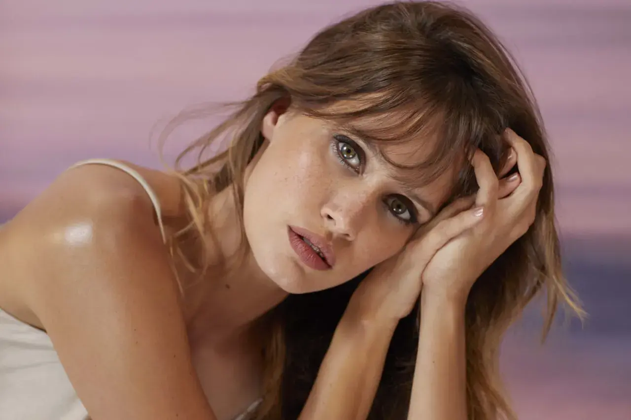 Portrait d'une jeune femme aux yeux verts perçants, sa frange effilée encadrant son visage. Elle pose ses mains près de ses joues, le regard intense.