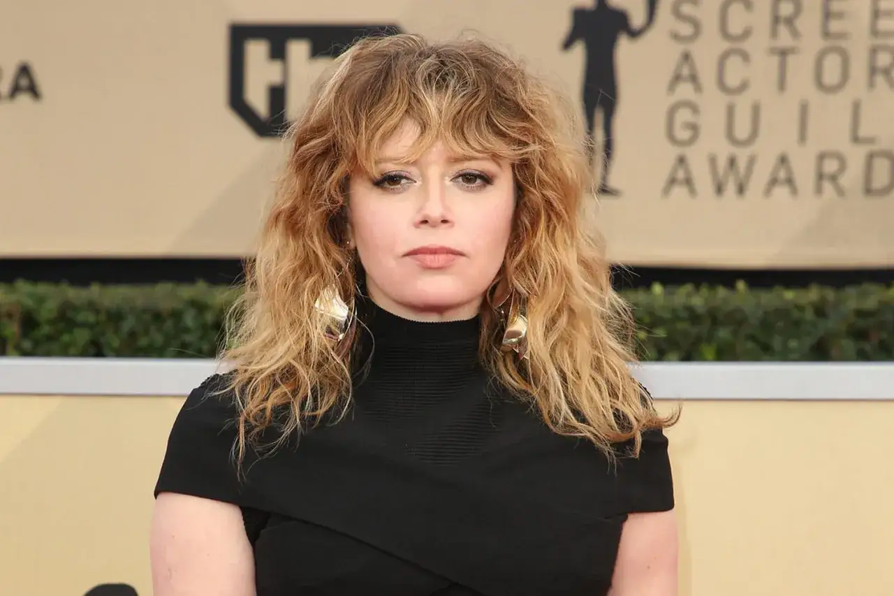 Natasha Lyonne, avec sa chevelure blonde ondul&eacute;e et sa frange effil&eacute;e, porte une robe noire et de grandes boucles d'oreilles dor&eacute;es.