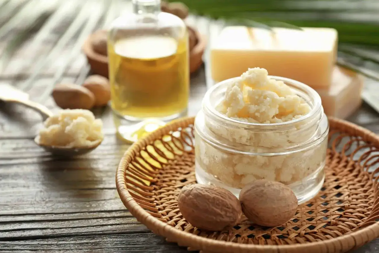 Un pot de beurre de karit&eacute;, id&eacute;al pour les bienfaits cheveux, avec une cuill&egrave;re, une bouteille d'huile et des noix sur un plateau en osier.