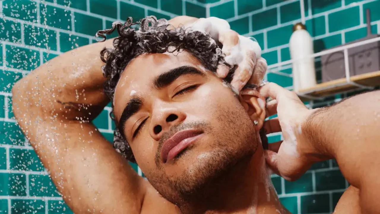 Un homme aux cheveux boucl&eacute;s savoure le moment de comment bien laver ses cheveux sous la douche, la mousse onctueuse dans ses mains.