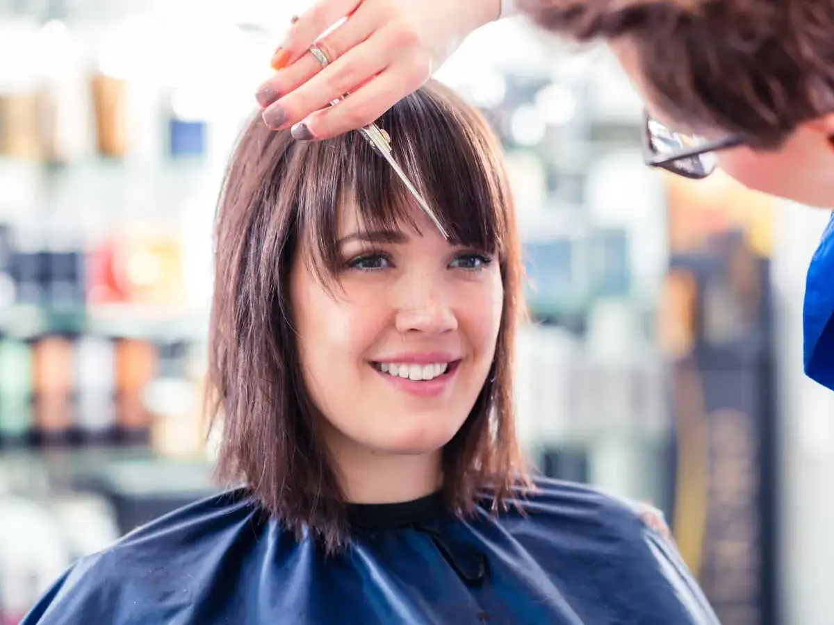 Une coiffeuse coupe la frange d'une cliente souriante. Le choix de quelle frange pour quel visage est crucial pour un look parfait.