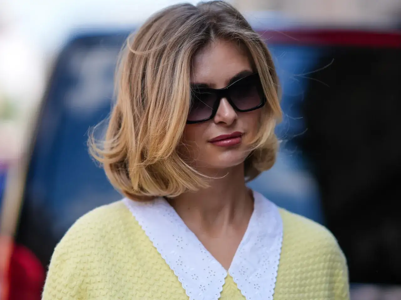 Une femme au brushing souple, lunettes de soleil oversize, porte un pull jaune pâle à col blanc brodé.
