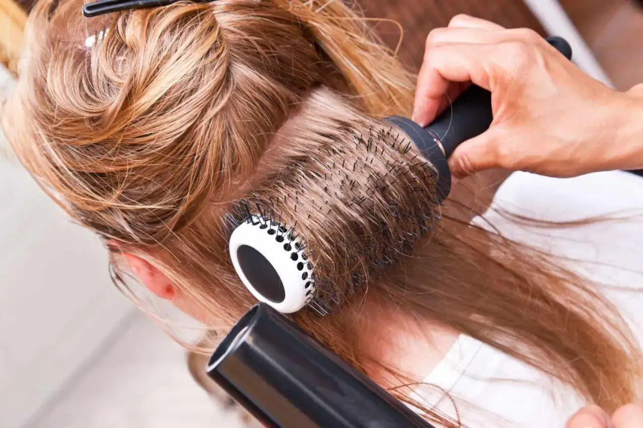 Une main coiffeuse r&eacute;alise un brushing sur des cheveux blonds avec une brosse ronde et un s&egrave;che-cheveux.
