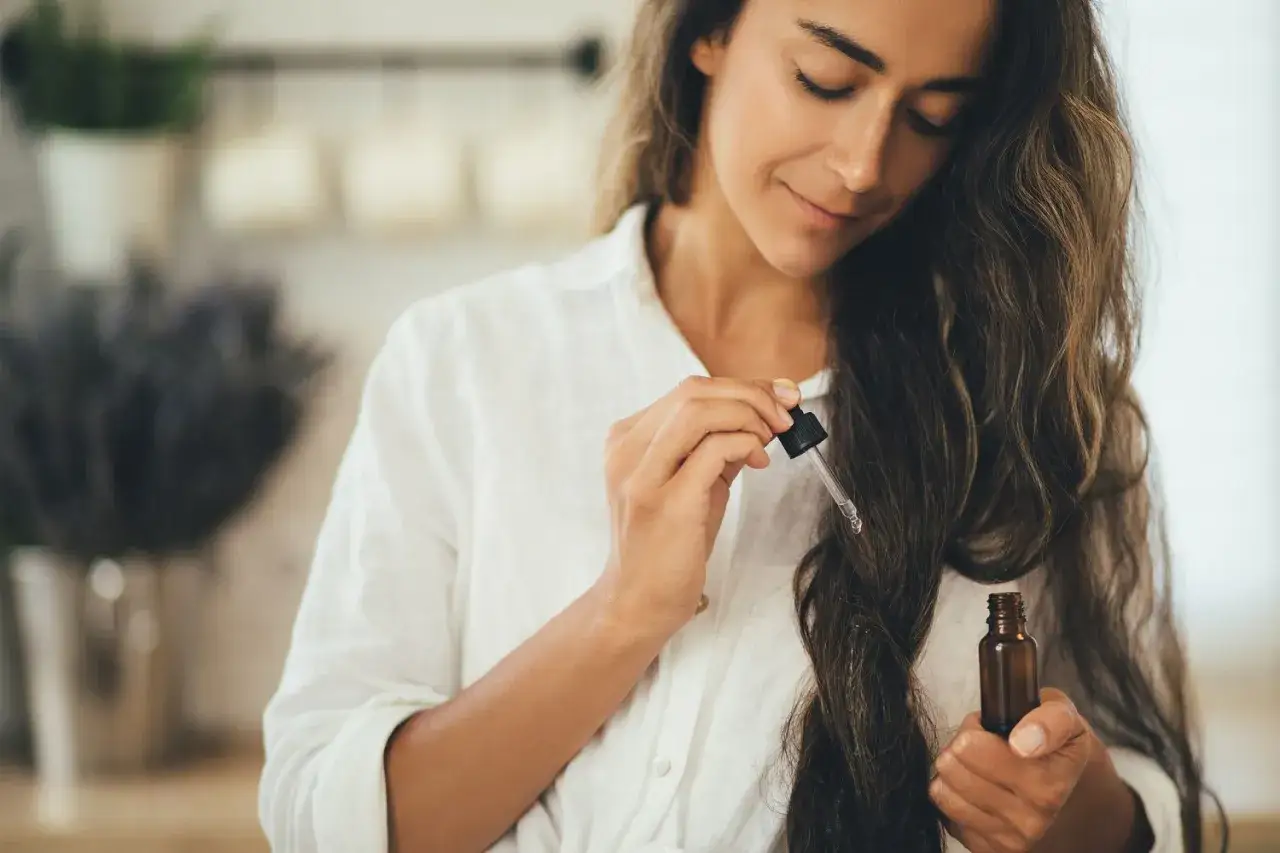 Une femme applique de l'huile dans les cheveux avec un compte-gouttes. Ses yeux sont fermés, elle savoure ce moment de soin.