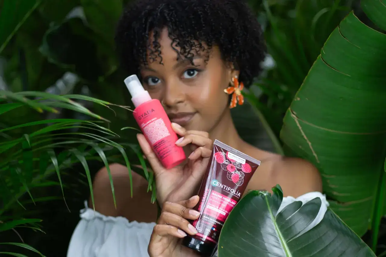 Une femme aux cheveux bouclés tient des produits capillaires, une solution pour le frisottis définition et un baume démêlant.