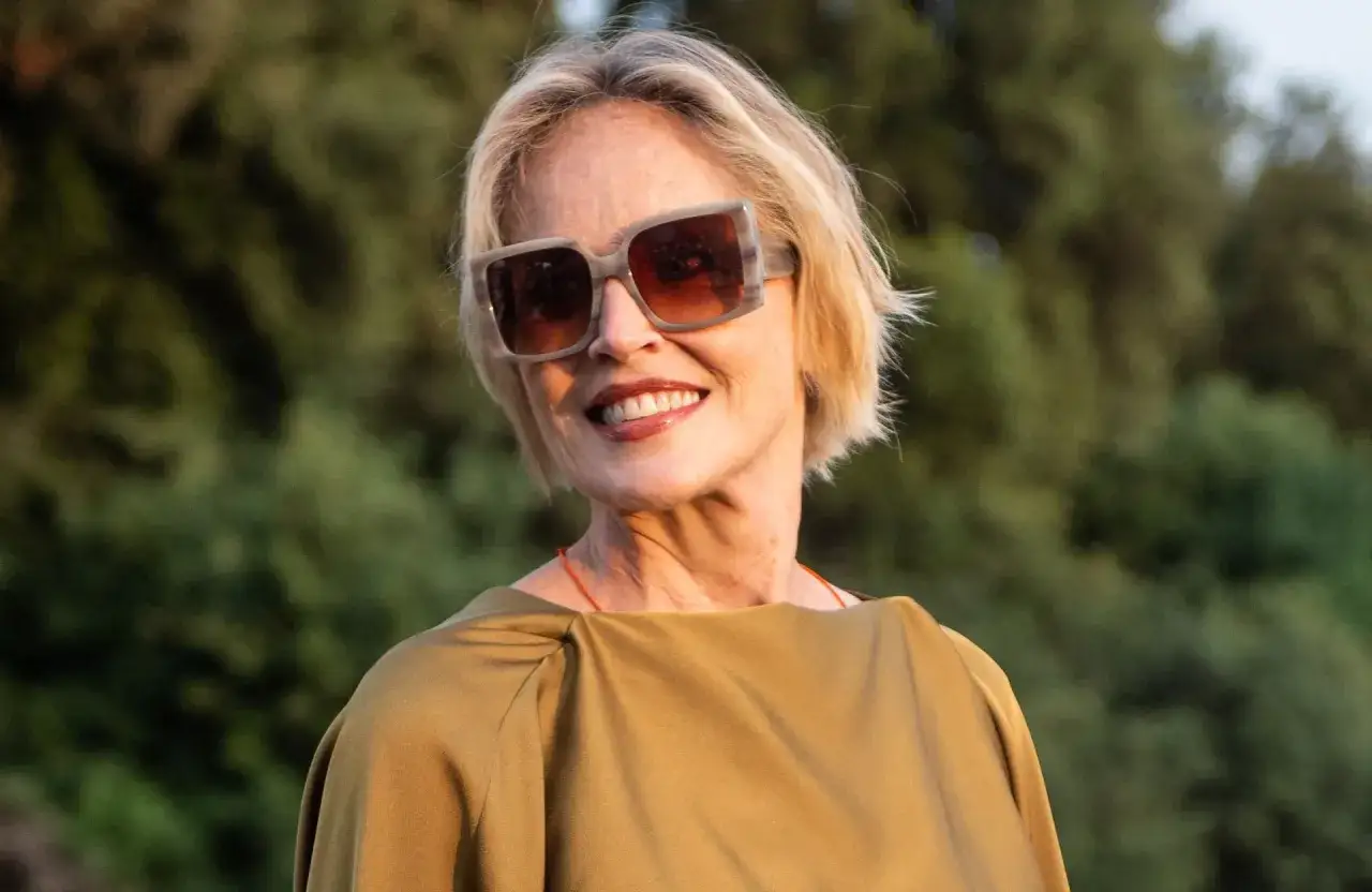Une femme d'environ 60 ans au visage ovale, avec une coupe de cheveux courte et blonde, porte de grandes lunettes de soleil.