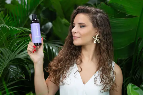 Une femme aux cheveux longs et bouclés tient un flacon de produit pour le soin des cheveux, entourée de verdure luxuriante.