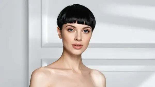 Une femme au regard intense arbore une coupe au bol audacieuse, ses cheveux noirs encadrant son visage aux traits fins.