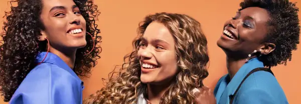 Trois femmes souriantes aux cheveux bouclés, chacune avec un style unique. Une inspiration pour savoir quelle frange pour quel visage.