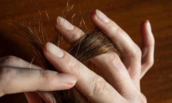 Des doigts effleurent une mèche de cheveux châtains, comme une **fourche à cheveux** naturelle, révélant des reflets dorés sous la lumière.
