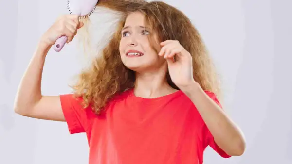 Une jeune fille aux cheveux mousseux et fins essaie de se brosser les cheveux, l'air désemparée.
