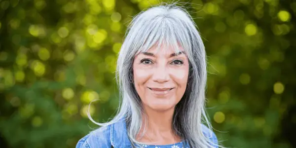 Une femme souriante aux cheveux gris argentés, un balayage subtil pour cacher les cheveux blancs, porte une veste en jean.
