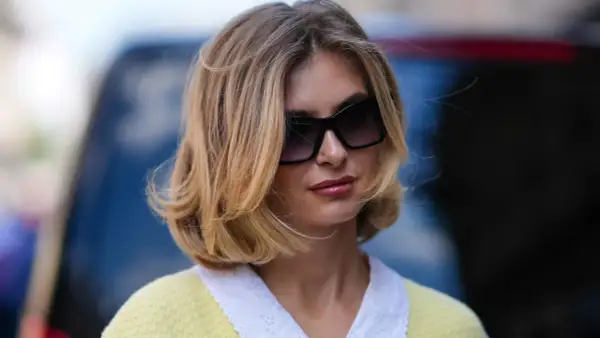Une femme avec une coupe cheveux mi long blonde, des lunettes de soleil oversize et un pull jaune à col blanc.