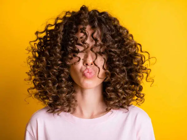 Une femme avec une magnifique coupe cheveux ondulés femme envoie un bisou, ses boucles brunes encadrant son visage sur fond jaune vif.