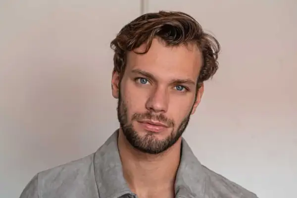 Un homme aux yeux bleus perçants arbore une coupe homme cheveux raides, légèrement ondulés sur le dessus, et une barbe courte.