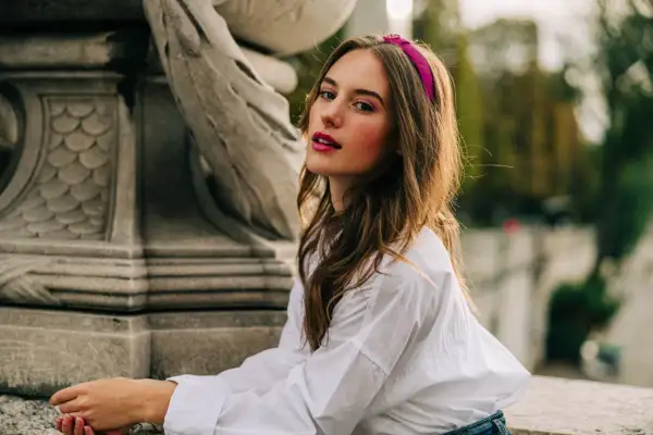 Jeune femme avec une coiffure avec serre tête fuchsia, chemise blanche, assise près d'une sculpture.