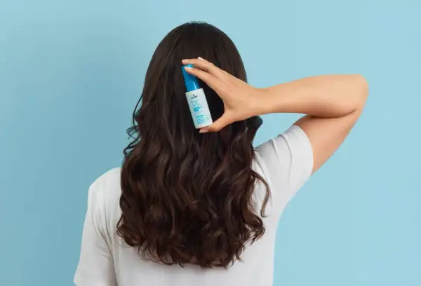 Une main tient un flacon de soin capillaire sur des cheveux bruns ondulés, pour lutter contre les cheveux déshydratés.