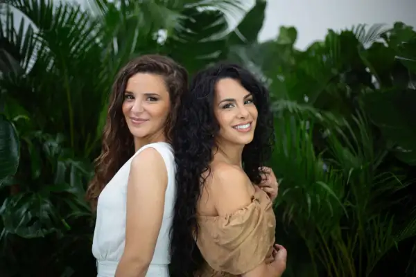 Deux femmes souriantes se tiennent dos à dos devant un feuillage luxuriant. L'une a des cheveux châtains ondulés, l'autre des cheveux noirs et bouclés, une magnifique chevelure mousseuse.