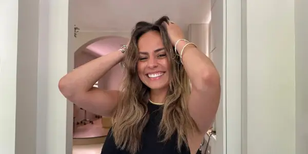 Une femme souriante avec des cheveux châtains ondulés, elle joue avec ses cheveux pour boucler cheveux. Elle porte des bracelets dorés.