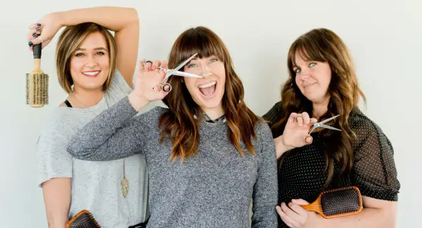 Trois femmes souriantes tiennent des ciseaux et des brosses. L'une d'elles joue avec ses ciseaux, comme pour savoir quand couper ses cheveux pour qu'ils poussent plus vite.